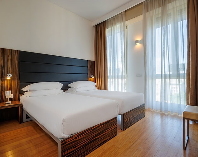 A modern hotel room with two single beds, white bedding, wooden floors, bedside tables with lamps, and large windows covered by sheer curtains letting in natural light.