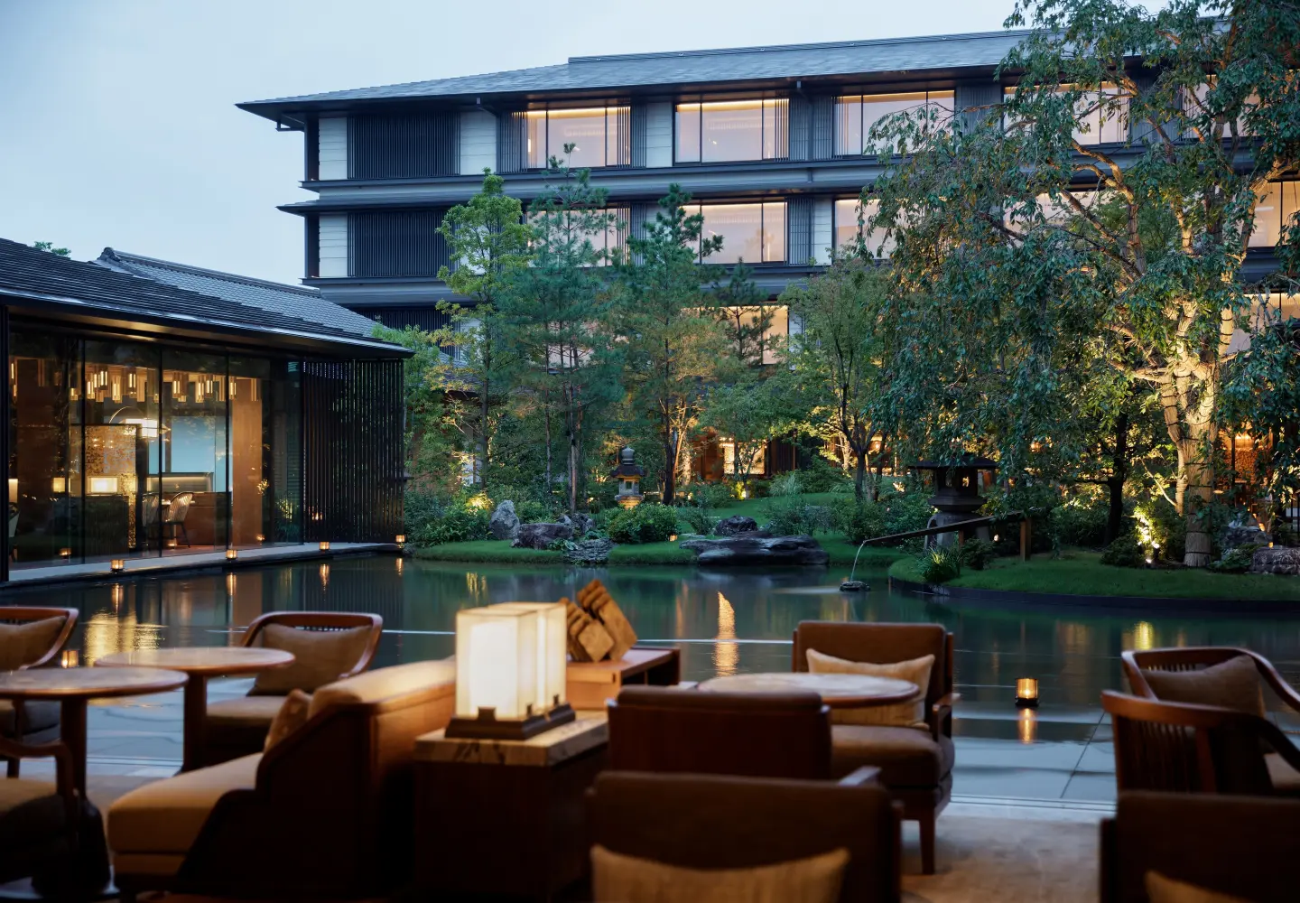 Outdoor seating area with chairs and tables overlooks a tranquil pond surrounded by trees and greenery. In the background, a modern multi-story building with large windows is softly illuminated at dusk.