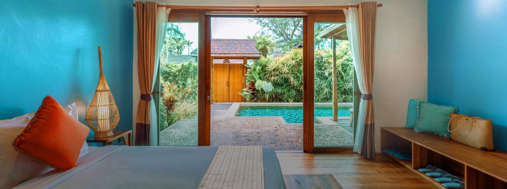 A bright, tropical bedroom with turquoise walls and wood floors features a bed with orange and blue pillows, large glass doors opening to a private pool, lush greenery, and outdoor seating.