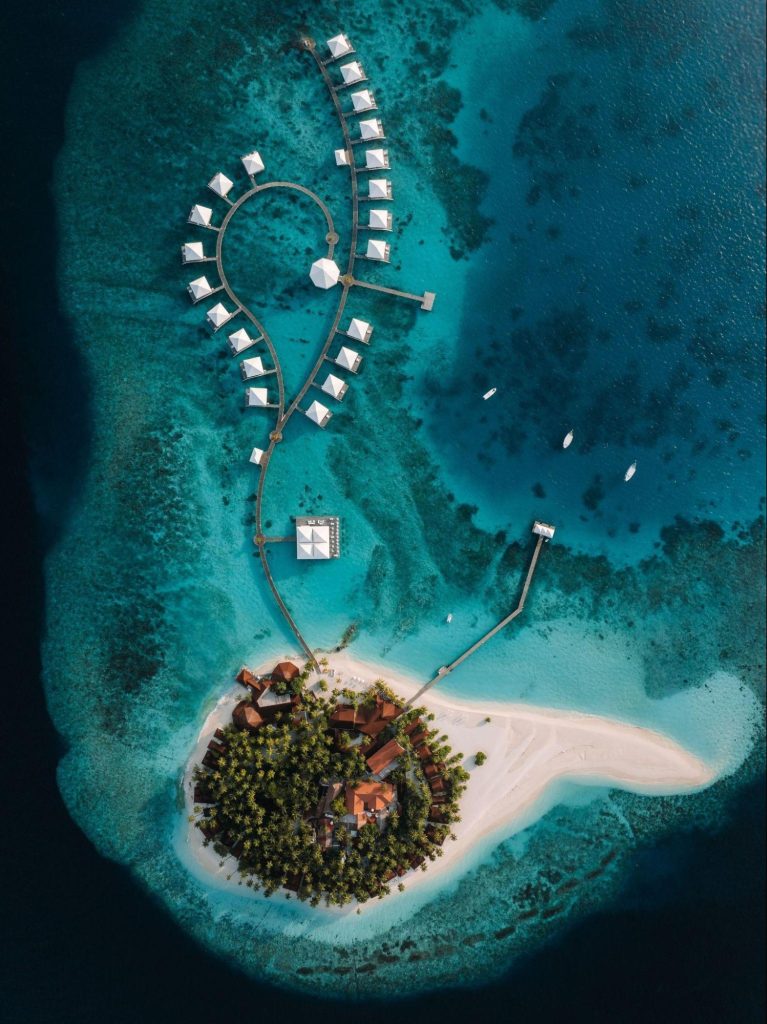 Aerial view of a tropical island resort with overwater bungalows, white sandy beaches, turquoise waters, palm trees, and a dock extending into the sea. Small boats float nearby in the surrounding lagoon.