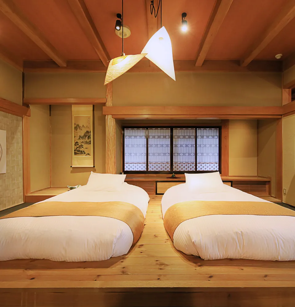 A cozy Japanese-style bedroom with two low twin beds, each with white linens and tan throws, wooden floors, shoji-style window, a modern pendant light, and traditional décor elements.