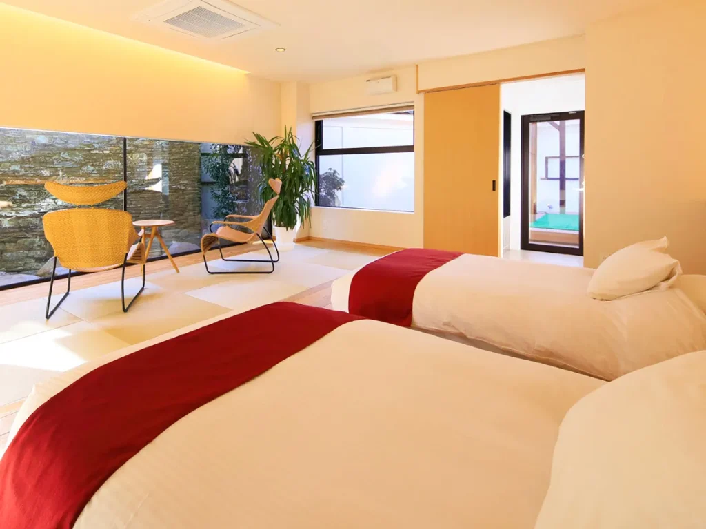 A bright, modern bedroom with two twin beds topped with white linens and red throws, a seating area with woven chairs and a small table, large windows, and minimalistic decor.