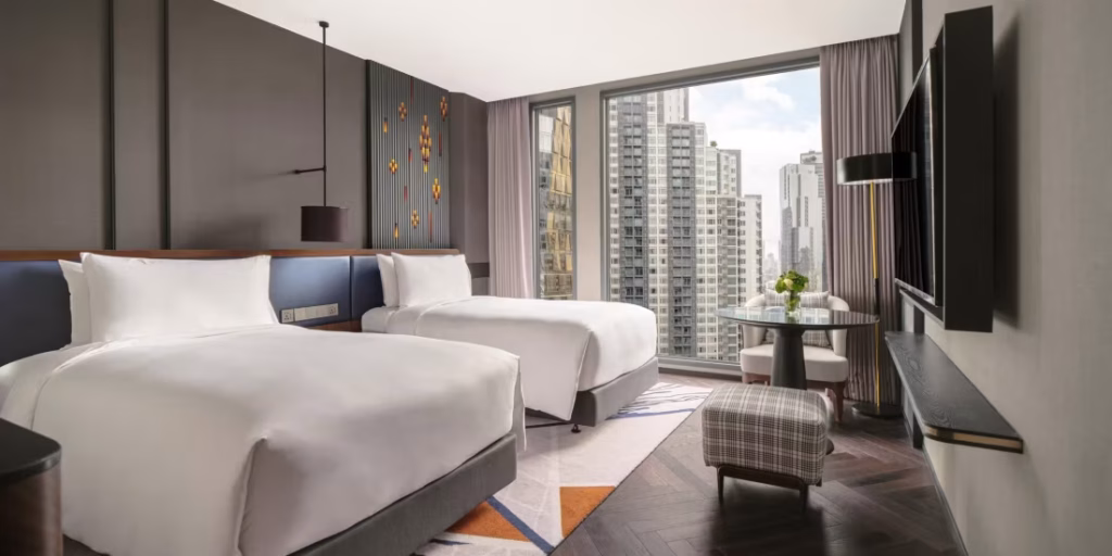 A modern hotel room with two double beds, a large window showing city buildings, a small round table with chairs, a flat-screen TV on the wall, and contemporary décor in neutral tones.