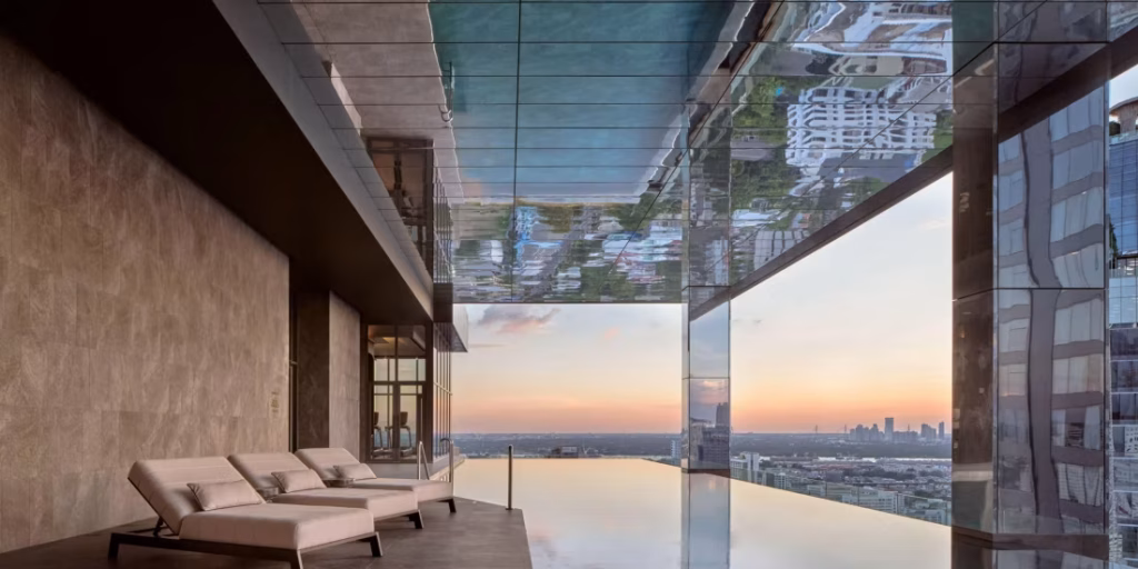 A modern rooftop infinity pool with mirrored ceiling and glass walls overlooks a city skyline at sunset. Three lounge chairs sit on the pool deck beside the water, creating a serene, luxurious atmosphere.