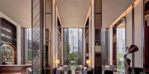 Modern hotel lobby with tall glass windows, high ceilings, and elegant vertical columns. Contemporary furniture and sculptures are arranged for seating, with city buildings and green trees visible outside. Warm lighting enhances the space.