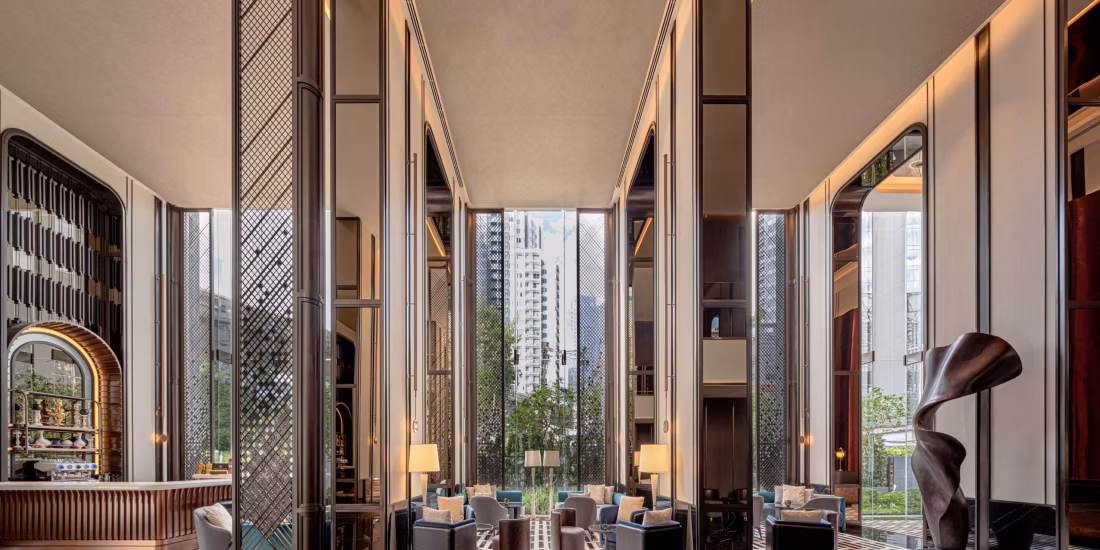 Modern hotel lobby with tall glass windows, high ceilings, and elegant vertical columns. Contemporary furniture and sculptures are arranged for seating, with city buildings and green trees visible outside. Warm lighting enhances the space.