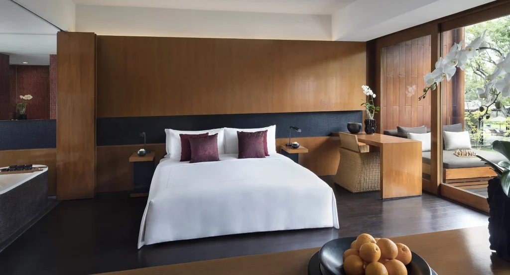 A modern hotel room with a large bed, white linens, and two purple cushions. Wooden walls, a desk with a chair, orchids, a bowl of oranges, and large windows with outdoor views complete the serene space.