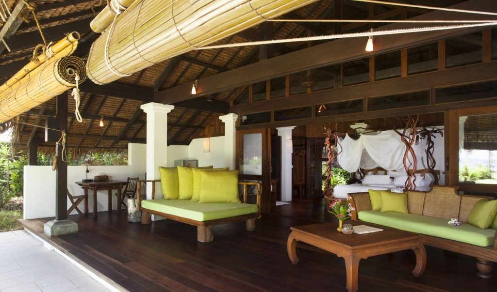 Open-air tropical villa with wooden floors, bamboo blinds, green cushioned seating, a wooden coffee table, and a cozy bedroom with white mosquito netting visible in the background. Lush greenery can be seen outside.