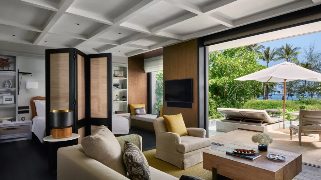 Spacious, modern hotel suite with neutral-toned furniture, an armchair, a sofa, a desk, and large sliding doors opening to a patio with lounge chairs, umbrella, greenery, and an ocean view in the background.