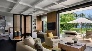 Spacious, modern hotel suite with neutral-toned furniture, an armchair, a sofa, a desk, and large sliding doors opening to a patio with lounge chairs, umbrella, greenery, and an ocean view in the background.
