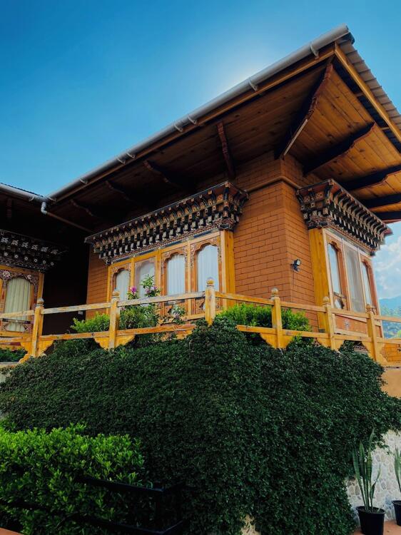 A traditional wooden house with ornate window frames, a sloped roof, and a balcony bordered by greenery and lush plants, set against a clear blue sky.