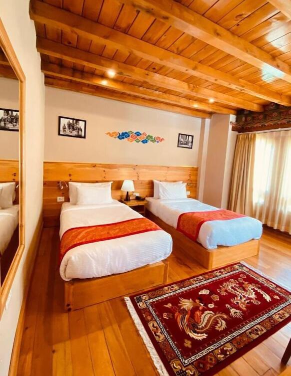 A cozy hotel room with wooden floors and ceiling, featuring two single beds with white bedding and red runners, a decorative rug, bedside table with lamp, wall art, and a large window with beige curtains.