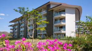 A modern multi-story apartment building with large balconies, surrounded by green trees and bright pink flowers in the foreground, under a clear blue sky.