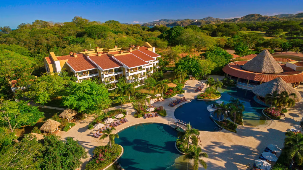 Aerial view of a resort surrounded by lush greenery, with a large pool, palm trees, lounging areas, thatched-roof structures, and buildings set against distant hills under a clear blue sky.