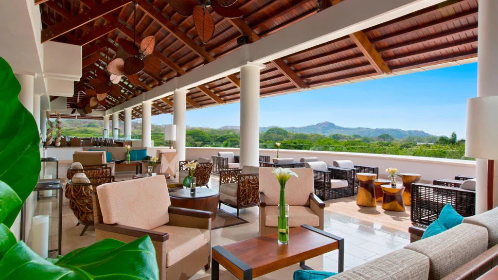 Open-air lounge with cushioned chairs and tables, tropical plants, and ceiling fans, overlooking lush green hills and distant mountains under a clear blue sky.