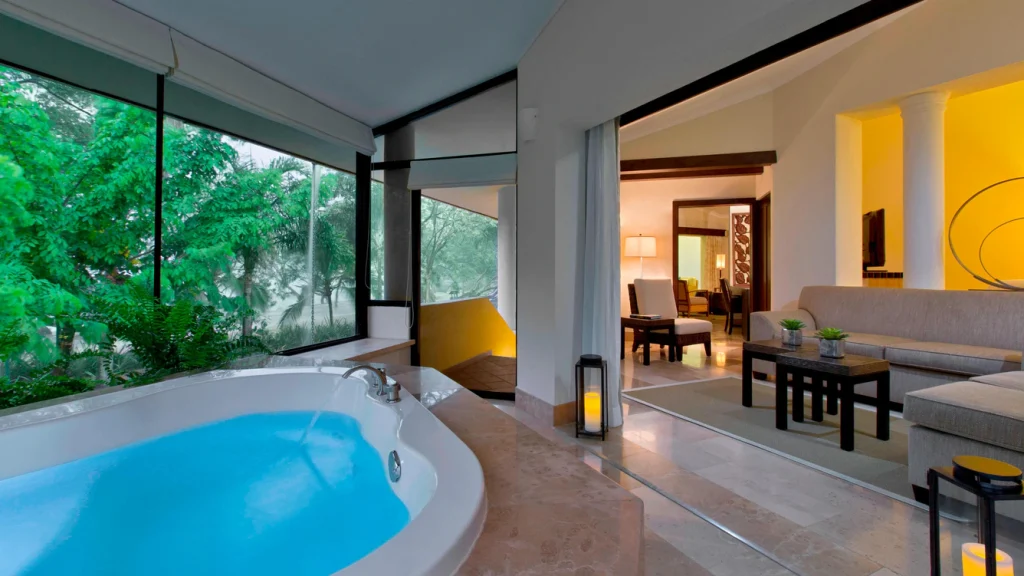 A luxurious bathroom with a round bathtub filled with water, large windows showing green trees, and an open view into a modern living area with a sofa, tables, and warm lighting.