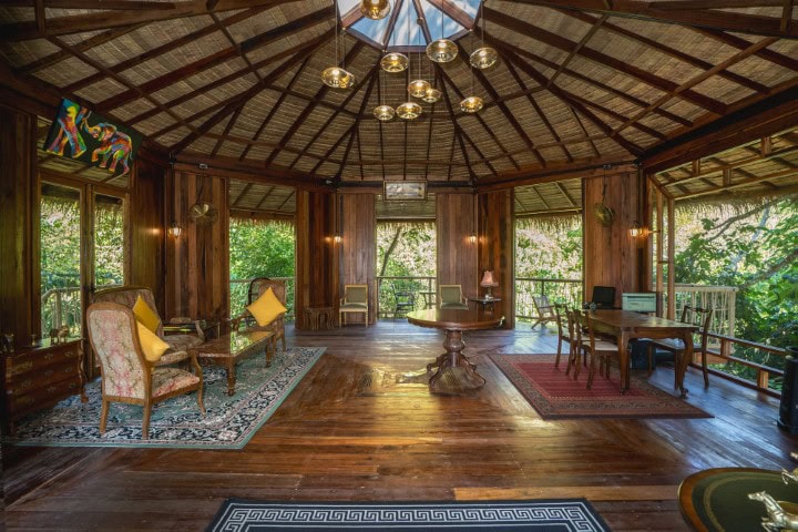 A spacious, octagonal wooden room with high thatched ceilings, large windows showcasing lush greenery, elegant chairs, rugs, a round table in the center, and warm natural light filling the space.
