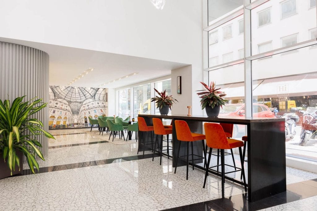 A modern, bright cafe with floor-to-ceiling windows, orange bar stools at a high counter, green chairs at tables, indoor plants, and a mural of a grand glass-domed building on the back wall.