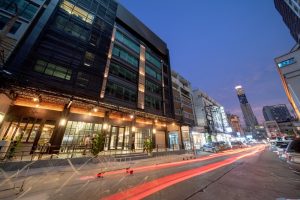 A city street at dusk with light trails from passing vehicles, modern buildings with illuminated windows, and a tall skyscraper visible in the distance under a blue evening sky.