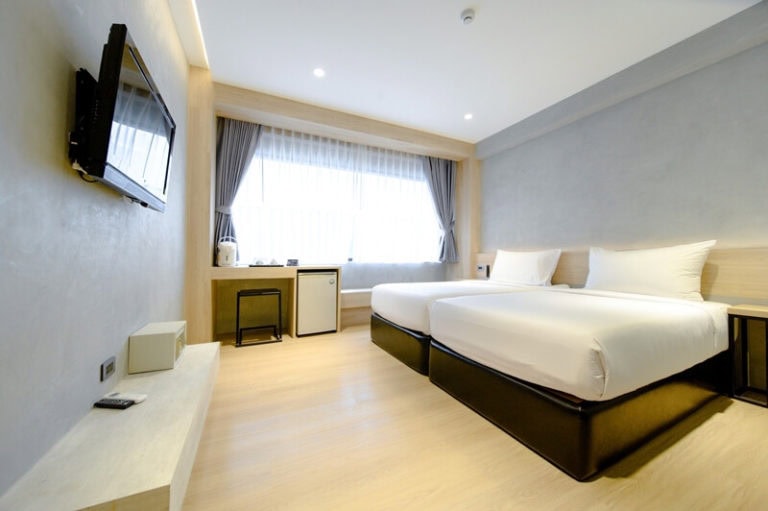 A modern hotel room with two neatly made single beds, a wall-mounted TV, a mini-fridge, and a large window with grey curtains letting in natural light. The decor is minimalist with light wood accents.