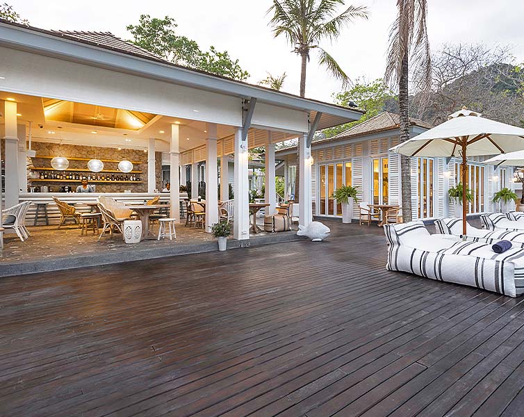 A spacious outdoor deck with lounge chairs and large umbrellas, adjacent to a modern open-air restaurant with wooden floors, stylish seating, and warm lighting, surrounded by palm trees and greenery.