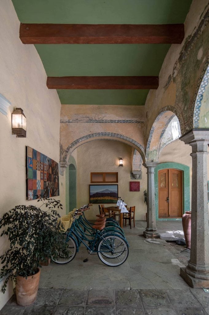 A rustic, covered courtyard with arched columns, green ceiling beams, two bicycles, potted plants, colorful wall art, and a small dining table near wooden doors. Natural light brightens the textured walls.