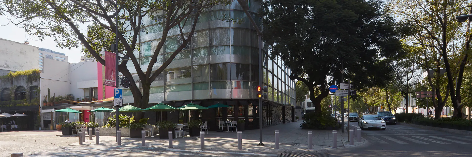 A modern glass building with green umbrellas at a street corner café, surrounded by trees and parked cars on a sunny day. A pedestrian crossing and street signs are visible.