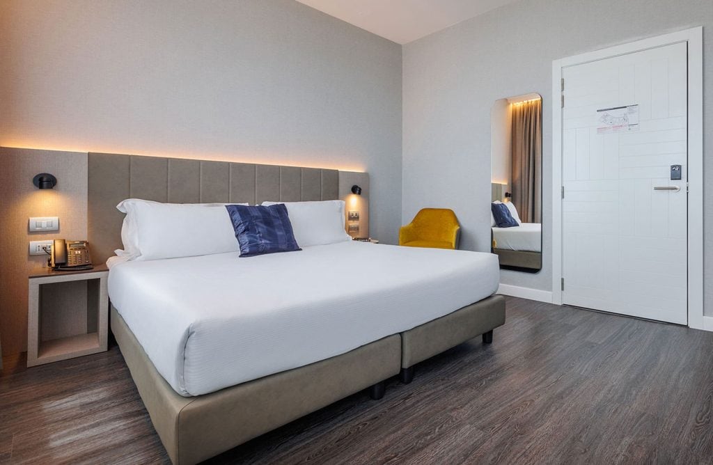 Modern hotel room with a large neatly made bed, a gray padded headboard, bedside tables, a yellow chair, a wall mirror, and a white door. The flooring is wood, and there is soft ambient lighting above the bed.