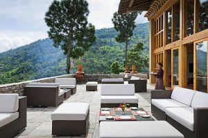 Outdoor terrace with modern white and brown patio furniture, set for a meal. A woman carries a tray near a building with large glass windows, overlooking lush green hills and tall trees.