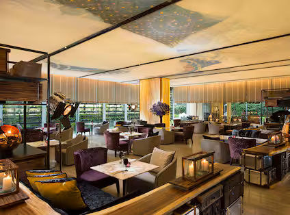 A modern, elegantly decorated restaurant with plush chairs, marble tables, warm lighting, large windows, and greenery visible outside. Decorative lamps and floral arrangements add to the cozy, upscale atmosphere.
