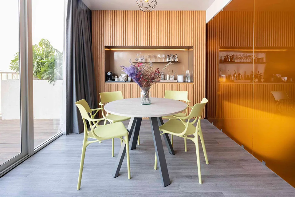 A modern dining area with a round table, four light yellow chairs, and a vase of dried flowers. Behind, a wooden bar with shelves and glassware; to the right, a reflective orange wall. Large windows let in natural light.