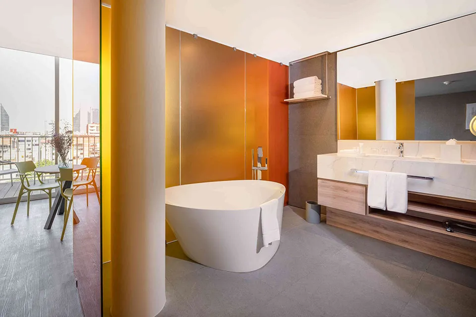 Modern bathroom with a freestanding white bathtub, large mirror, wooden vanity with white sinks and towels, frosted glass partition, and a view of a cityscape through floor-to-ceiling windows.