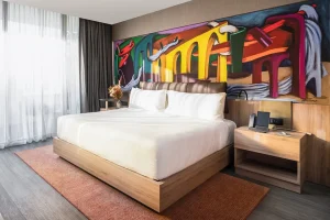 A modern hotel room with a large bed, white linens, wooden furniture, a colorful abstract mural above the headboard, a nightstand with a phone, and sunlight filtering through sheer curtains.