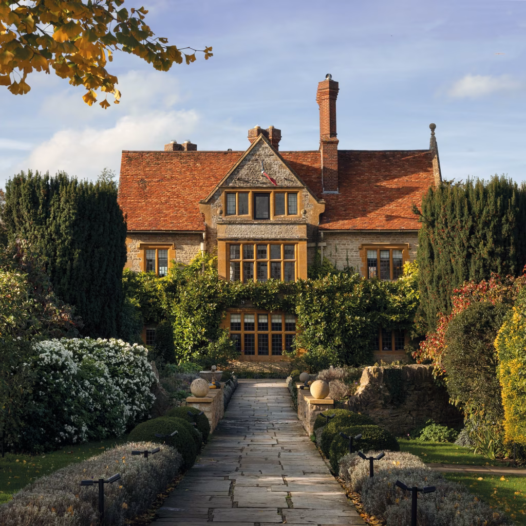 A large stone manor with red-tiled roof and tall chimneys, surrounded by lush gardens, hedges, and flowering bushes, under a clear blue sky with sunlight illuminating the building and autumn-colored trees.