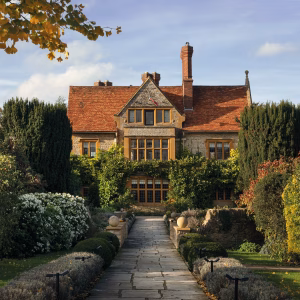 A large stone manor with red-tiled roof and tall chimneys, surrounded by lush gardens, hedges, and flowering bushes, under a clear blue sky with sunlight illuminating the building and autumn-colored trees.