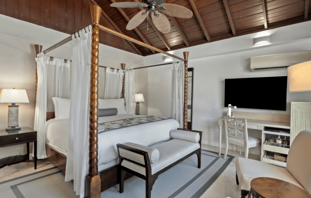 A stylish bedroom features a four-poster canopy bed with white linens, a cushioned bench at the foot, a flat-screen TV, a writing desk, wooden ceiling, and neutral-toned décor with modern and tropical touches.