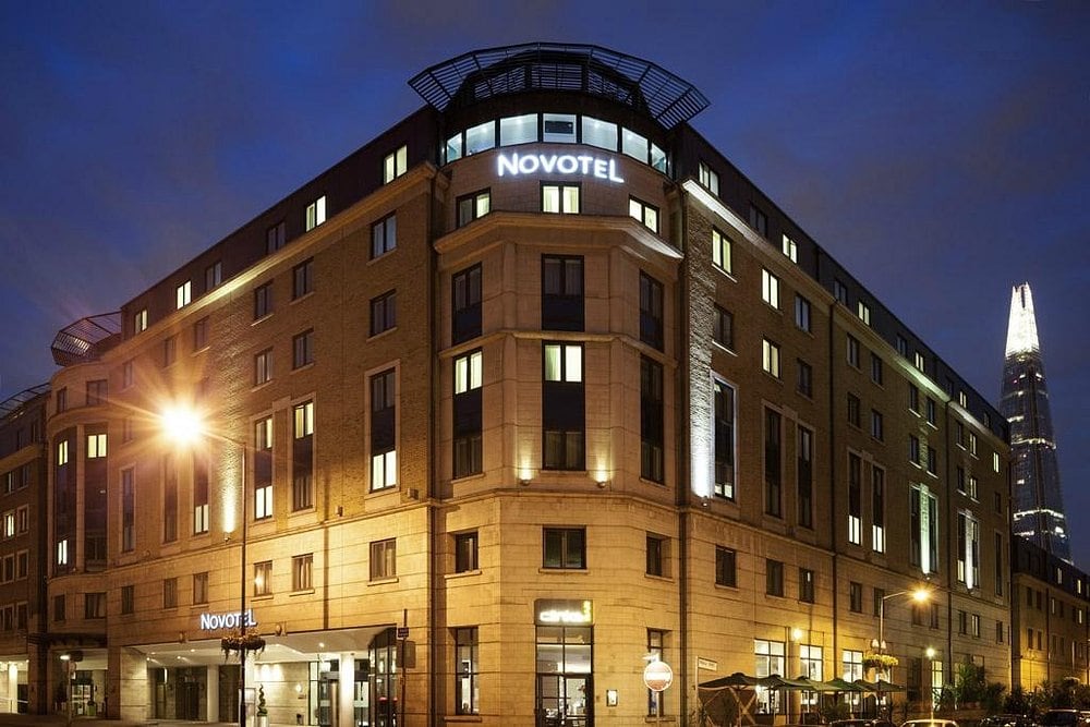 A modern, well-lit Novotel hotel building at dusk, with glowing windows and the Shard skyscraper visible in the background against a deep blue sky.