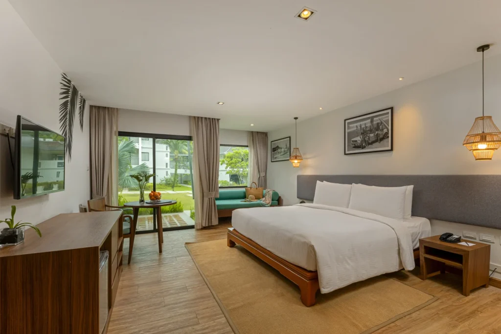A spacious hotel room with a large bed, a wooden desk, a green sofa by a window, wall art, hanging lights, and light-colored walls and floors, overlooking greenery outside.