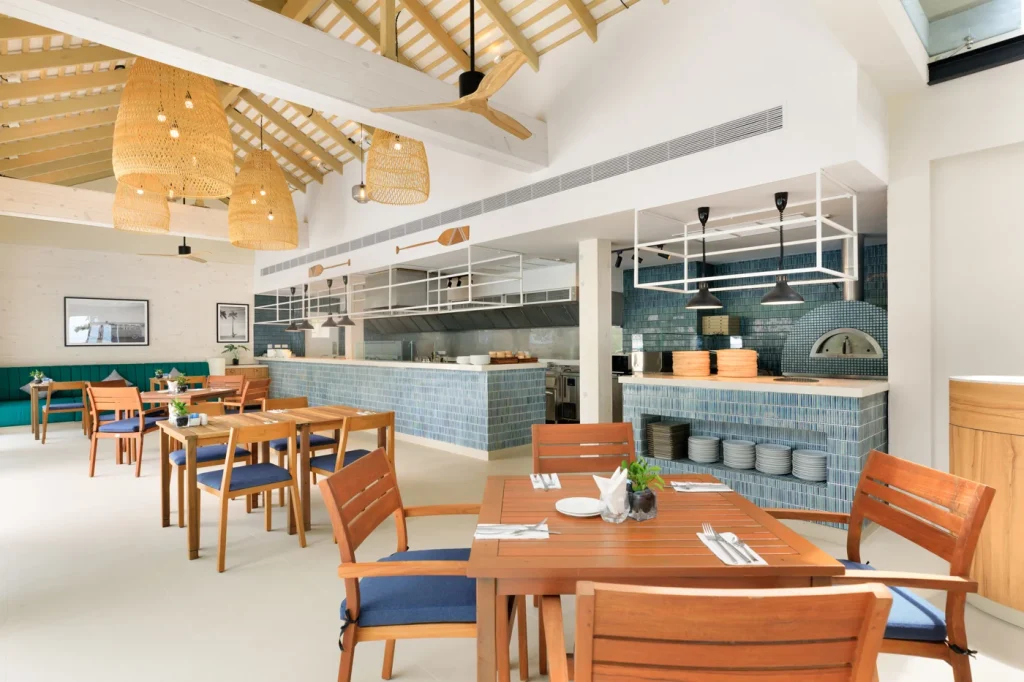 A bright, modern restaurant interior with wooden tables and blue cushioned chairs. Open kitchen with blue tile accents, wicker pendant lights, and decorative paddles on the wall. Natural light fills the airy space.