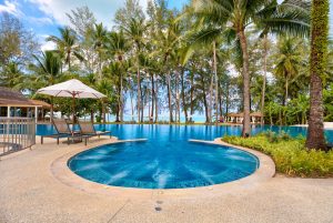 A round swimming pool with deck chairs and an umbrella sits among palm trees, with a view of the ocean and blue sky in the background.