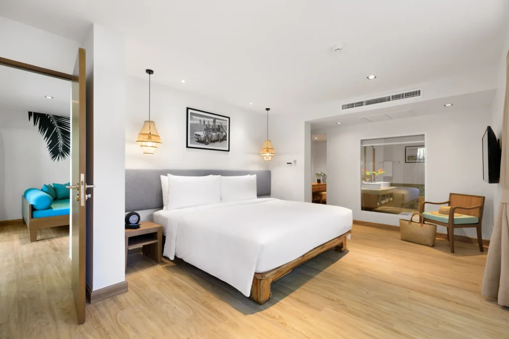 A modern hotel room with a large white bed, wooden floors, bedside tables, hanging lamps, a wall-mounted TV, a chair, and an open doorway leading to a sitting area with a blue sofa.