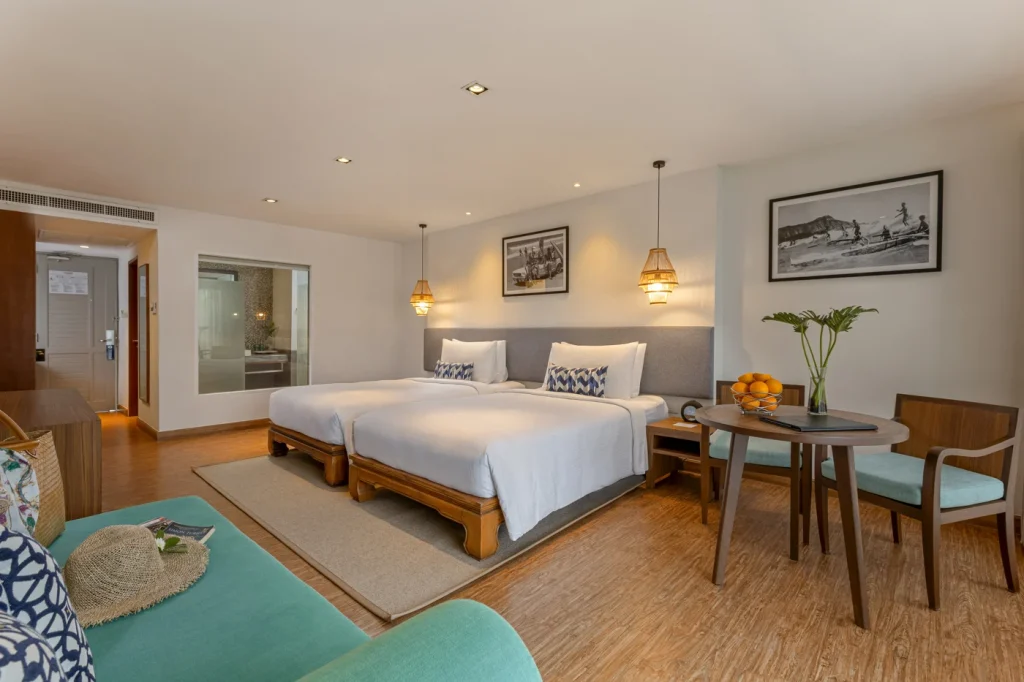 A spacious hotel room with two beds, white bedding, wooden furniture, a small round table with chairs, a bowl of fruit, and a vase with flowers. There are framed photos on the wall and a green sofa in the foreground.