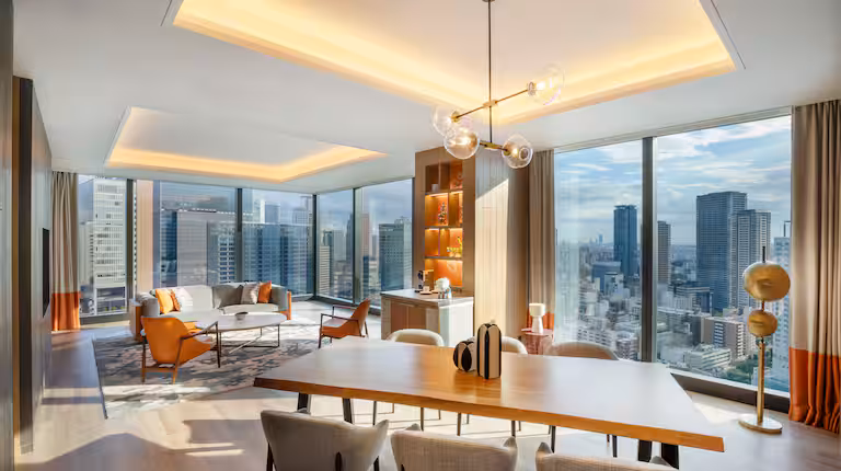 Modern high-rise apartment with floor-to-ceiling windows offering a city skyline view, featuring a dining table, contemporary chandelier, stylish lounge area, and warm, neutral decor with orange accents.