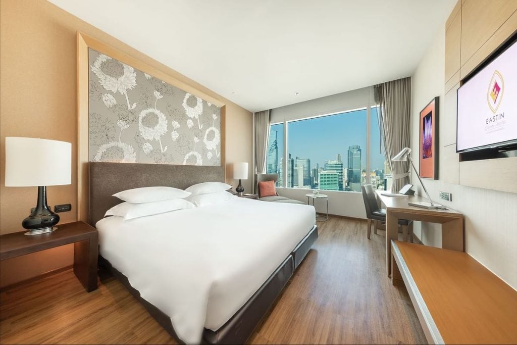Modern hotel room with a large bed, bedside tables and lamps, a desk and chair, wall-mounted TV, and a large window with a view of city skyscrapers under a clear blue sky.