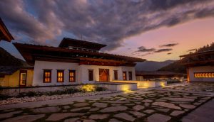 Traditional Bhutanese building with white walls, wooden roof, and glowing windows at sunset. Stone path and soft landscape lighting enhance the tranquil atmosphere under a dramatic, colorful sky.