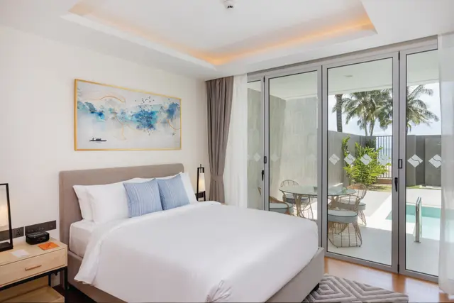 A modern bedroom with a neatly made bed, two blue accent pillows, abstract wall art, and floor-to-ceiling glass doors opening to a patio with outdoor seating, a pool, and palm trees outside.