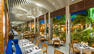 A stylish open-air restaurant with wooden tables and chairs, white tablecloths, modern decor, and lush greenery. The ceiling features unique sculpted panels, and palm trees are visible outside under evening lighting.