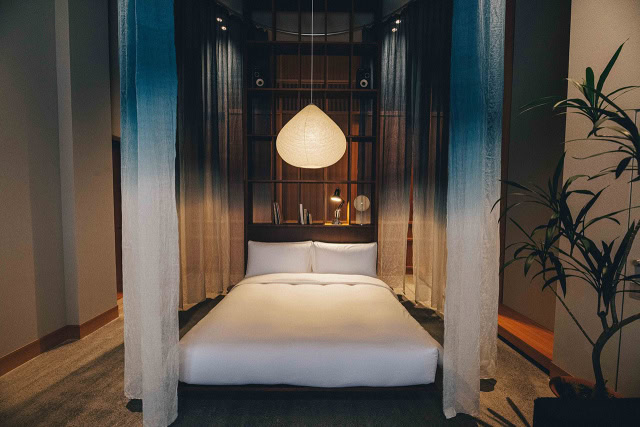A modern bedroom with a four-poster bed draped in sheer blue and white curtains, a large pendant light above, a wooden headboard with shelves, and a tall green plant on the right.