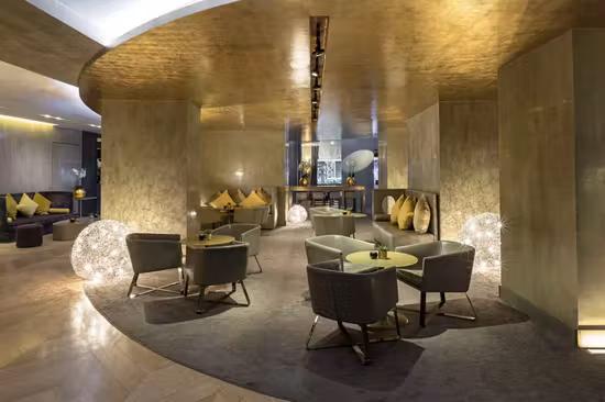 A modern lounge area with round tables, gray armchairs, yellow cushions, and large decorative light spheres, set against gold-toned walls and ceiling with soft, ambient lighting.