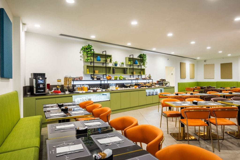 A modern dining area with orange chairs, green cushioned benches, and neatly set tables. A buffet counter at the back offers drinks and food, with shelves above displaying plants and decorative items. The space is brightly lit.